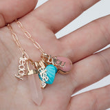 A hand holding an Element Short Link Chain with several charms, including Petite Nameplate Pendants 'bijou', 'teddy' and 'elise', an Etched Turquoise Heart Charm, a Balance Crystal Charm, an Engraved Heart Charm and an Engraved Heart Charm. 
