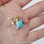 A hand holding an Element Short Link Chain with several charms, including Petite Nameplate Pendants 'bijou', 'teddy' and 'elise', an Etched Turquoise Heart Charm, a Balance Crystal Charm, an Engraved Heart Charm and an Engraved Heart Charm. 