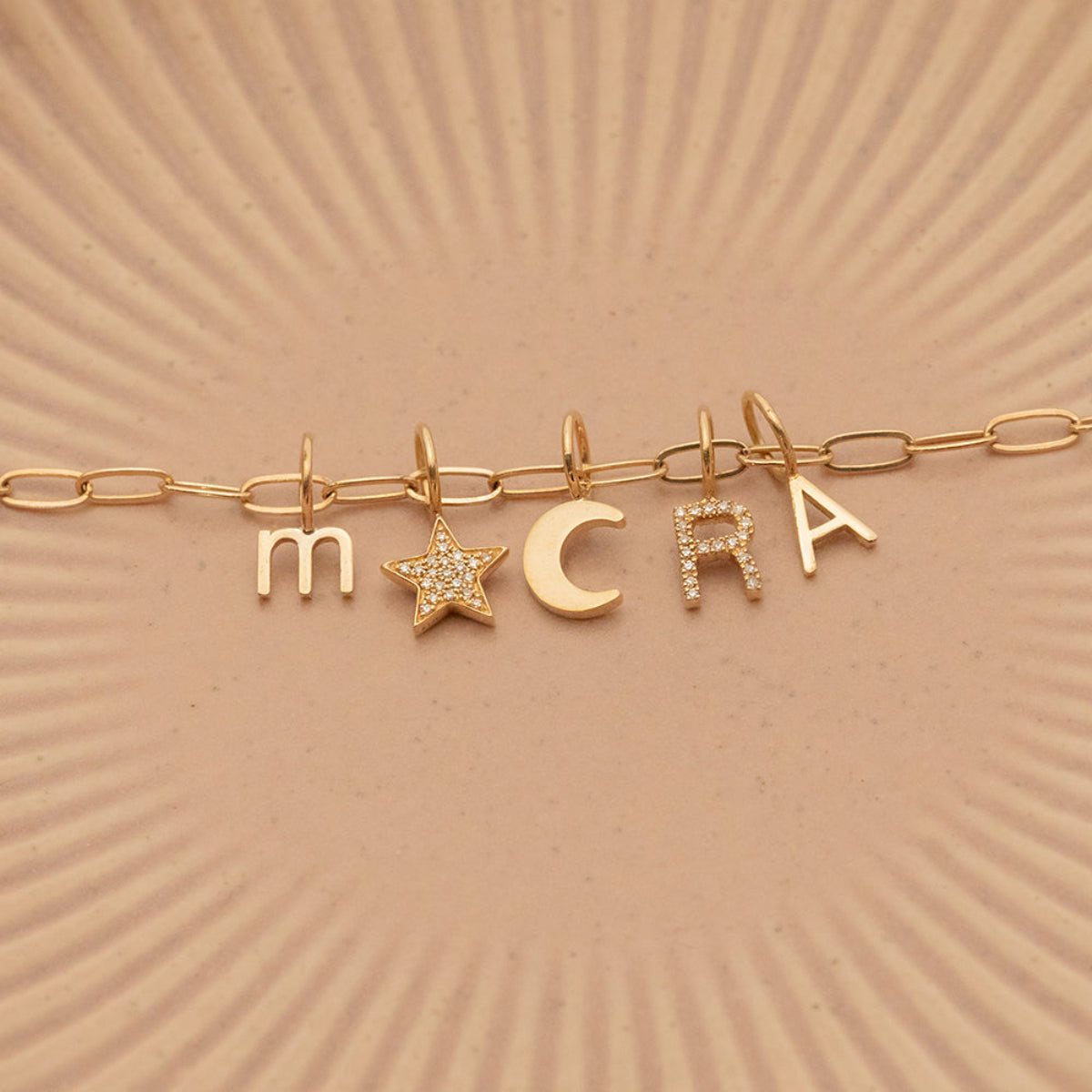 A Petite Letter Charm in m, a Gold Letter Charm in A, a Pavé Letter Charm in R, and Charm Pendants in Moon and Pavé Star on a Link Chain, all in yellow gold, on a beige background. 