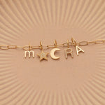 A Petite Letter Charm in m, a Gold Letter Charm in A, a Pavé Letter Charm in R, and Charm Pendants in Moon and Pavé Star on a Link Chain, all in yellow gold, on a beige background. 