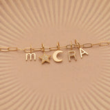 A Petite Letter Charm in m, a Gold Letter Charm in A, a Pavé Letter Charm in R, and Charm Pendants in Moon and Pavé Star on a Link Chain, all in yellow gold, on a beige background. 