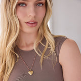 A blonde woman wearing The Peony Bouquet Necklace with a Diamond Heart Locket Charm and a brown top against a white background. 