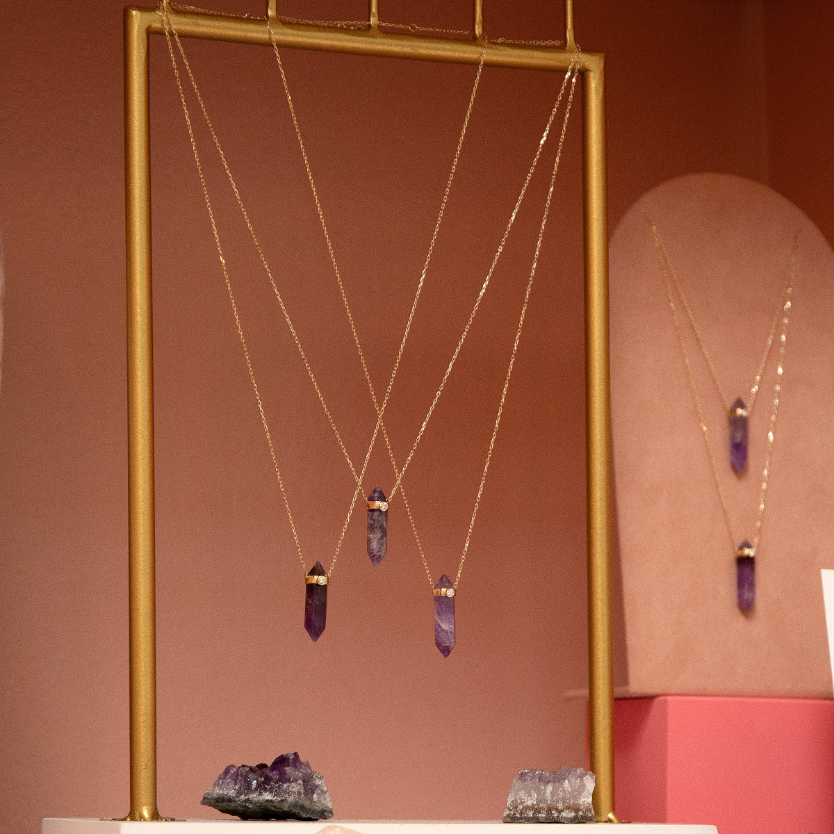 Several The Healing Retreat Necklaces on gold and pink stands with amethyst crystals against a light brown background. 