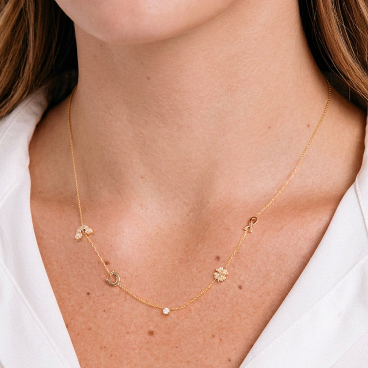 Gold necklace with rainblow , horseshoe, clover, and ribbin charms  on a person wearing a white shirt.