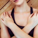A close-up of a woman with her hands on her shoulders, wearing a 14k Gold Asymmetrical Number Necklace - 2 in rose gold and various gold rings, including a Birthstone Ring, a Geo Stacking Ring, a 14K Gold Pavé Petal Ring and a Pave Geo Ring - Black Diamond, styled in a dark purple top. 