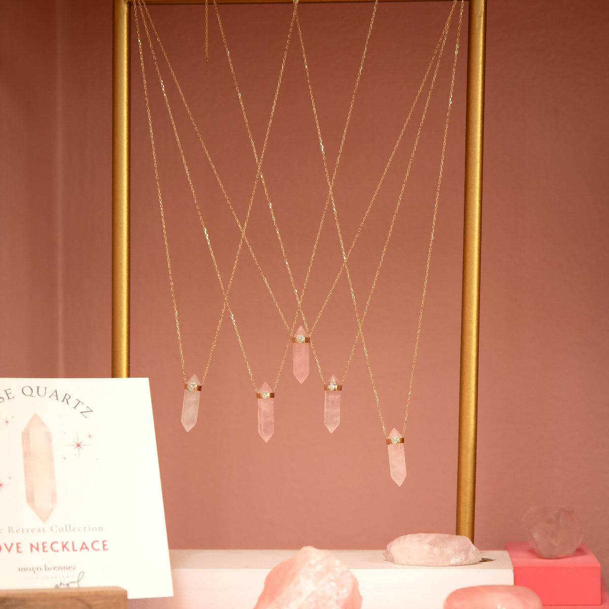 Several The Love Retreat Necklaces on a gold stand with rose quartz stones, against a pinky brown background. 