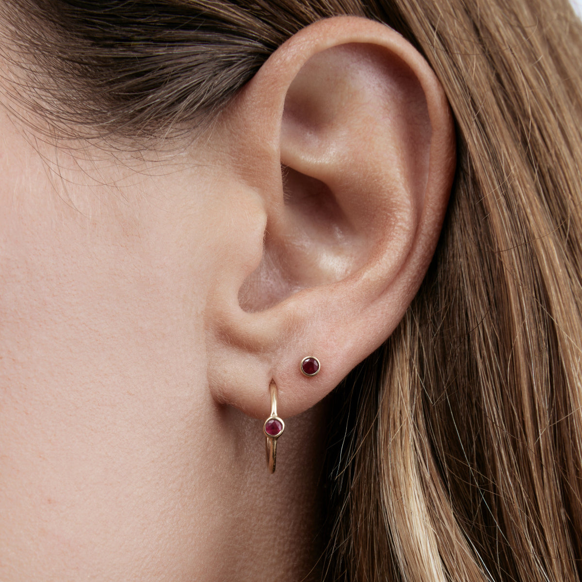 Close-up of a Birthstone Stud and Birthstone Hoop - Ruby (July) in a woman's ear,  with her brown hair visible. 