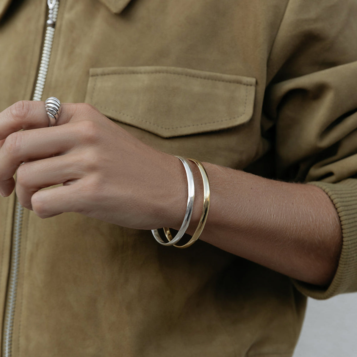 A close-up of an arm wearing two For Keeps Bangle, one in gold and one in silver and a white gold Etched Paris Ring, styled in a khaki jacket. 