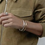 A close-up of an arm wearing two For Keeps Bangle, one in gold and one in silver and a white gold Etched Paris Ring, styled in a khaki jacket. 