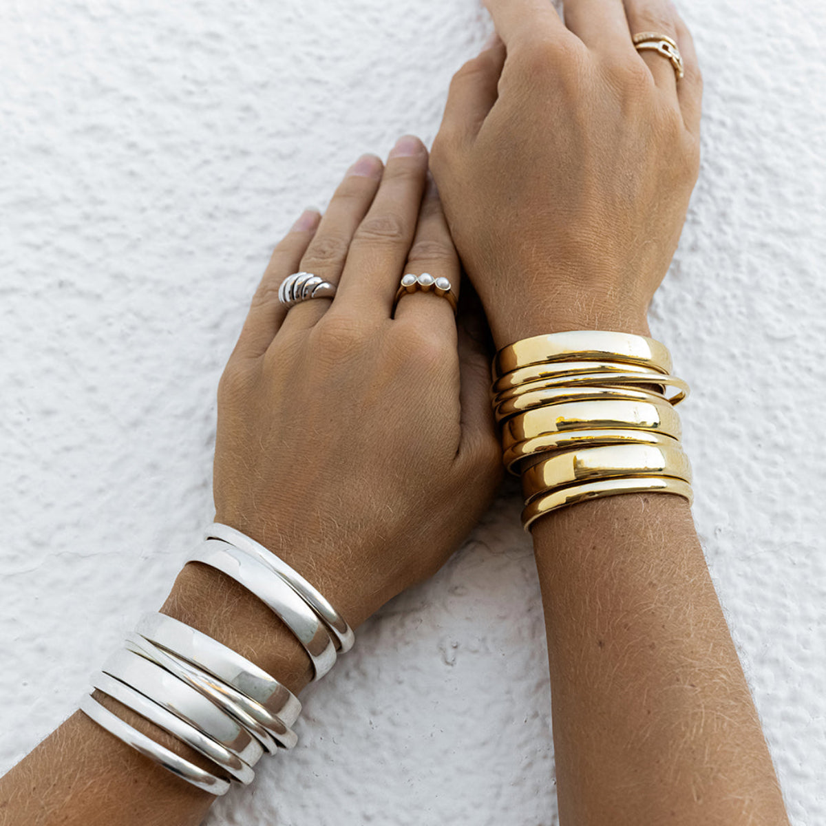 A close-up of arms wearing stacked Rendezvous Cuffs, Plus One Cuffs and All In Cuffs, one arm in silver and the other in gold, an Etched Paris Ring, a Tres Pearl Ring and a C'est Moi Link Ring against a textured white background.