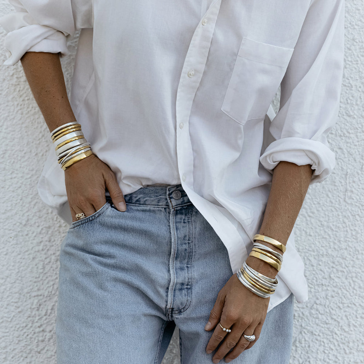 A person wearing silver and gold stacked Rendezvous Cuffs, Plus One Cuffs and All In Cuffs on both arms, an Etched Paris Ring, a Tres Pearl Ring, a C’est Moi Link Ring and an Open Diamond Ring, styled in a white shirt and light blue jeans.