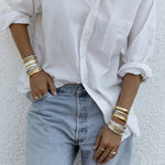 A person wearing silver and gold stacked Rendezvous Cuffs, Plus One Cuffs and All In Cuffs on both arms, an Etched Paris Ring, a Tres Pearl Ring, a C’est Moi Link Ring and an Open Diamond Ring, styled in a white shirt and light blue jeans.