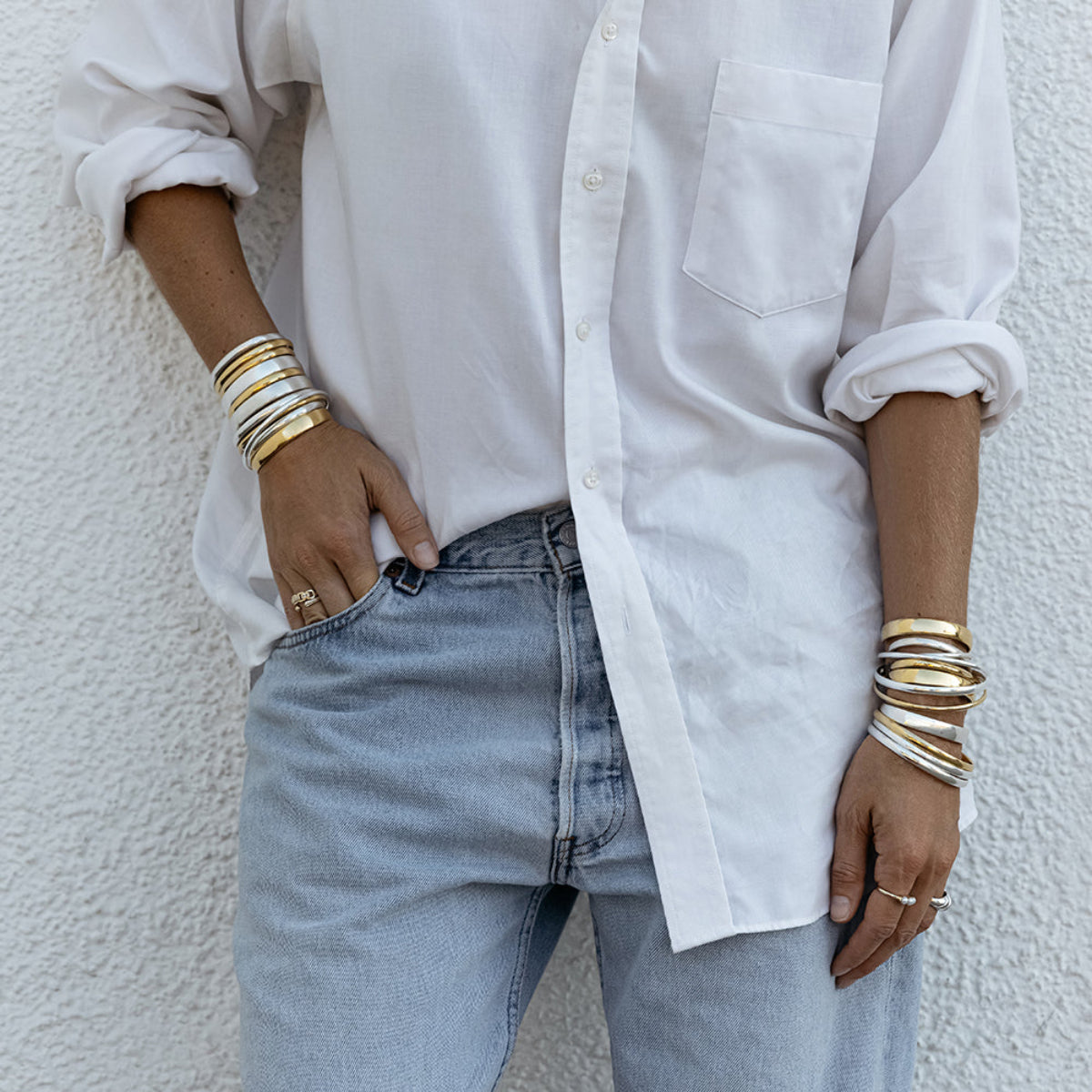 A person wearing silver and gold stacked Rendezvous Cuffs, Plus One Cuffs, All In Cuffs and Flirt Bangles on both arms, an Etched Paris Ring, a Tres Pearl Ring, a C’est Moi Link Ring and an Open Diamond Ring, styled in a white shirt and light blue jeans.