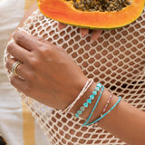 A hand wearing several Endless Summer Bracelets (Pink Sands, Turqs + Caicos, Coral Reef and Sea + Sky) and Bouquet Rings, holding a papaya in the other hand, wearing cream mesh clothing as the background.