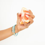 A hand wearing several Endless Summer Bracelets (Coral Reef, Sea + Sky, Pink Sands and Turqs + Caicos) holding a halved grapefruit against a white background. 
