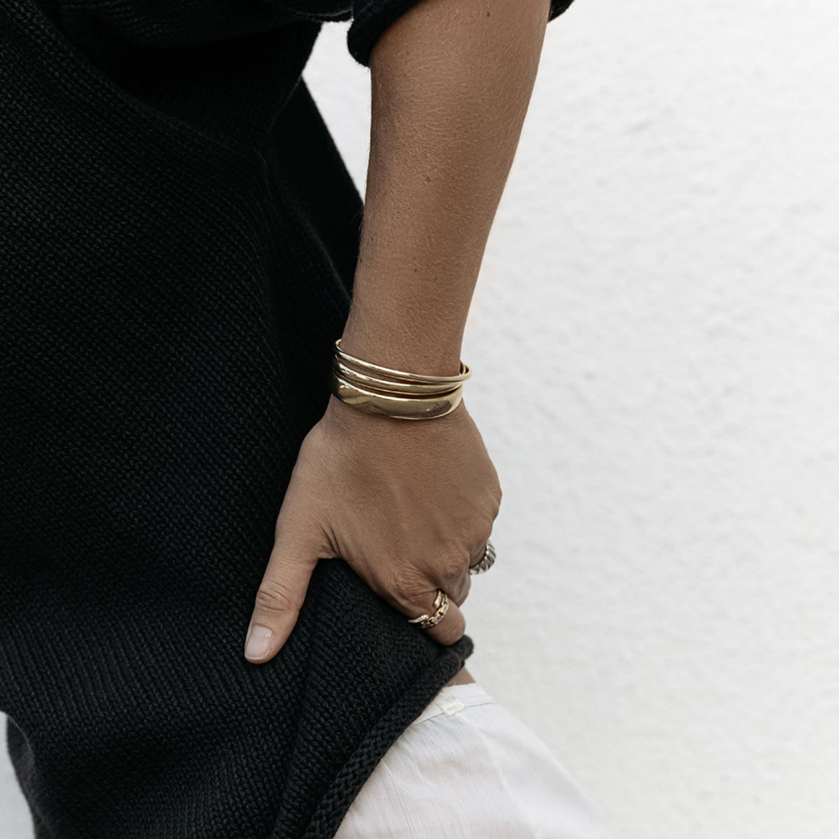 Close-up of a woman’s hand wearing two yellow gold Rendezvous Cuffs, an All In Cuff and rings, resting on her hip, styled in a black knit against a white background.