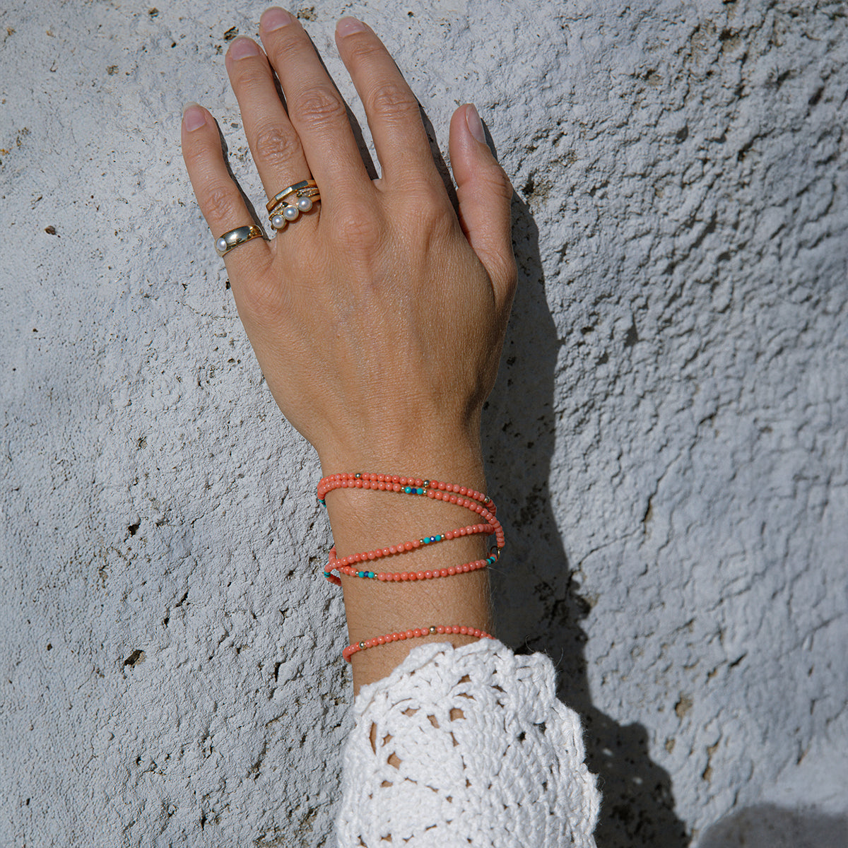 A hand wearing a Perla Pinky Ring, Tres Pearl Ring, an Open Diamond Ring, a Tenfold Ring and an Endless Summer Necklace - Calypso and an Endless Summer Bracelet - Mykonos with a light stone background. 