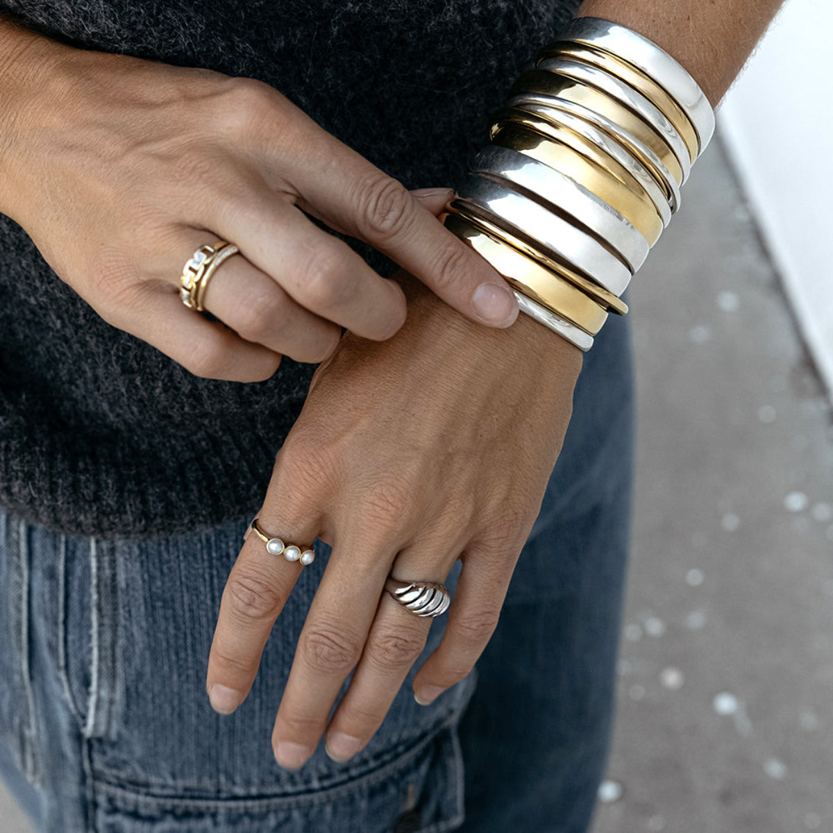 A pair of hands wearing several silver and gold All In Cuffs, Plus One Cuffs and Rendezvous Cuffs, along with a Tres Pearl Ring, Etched Paris Ring and C’est Moi Link Ring, styled with a grey knit top and denim against a softly blurred background.