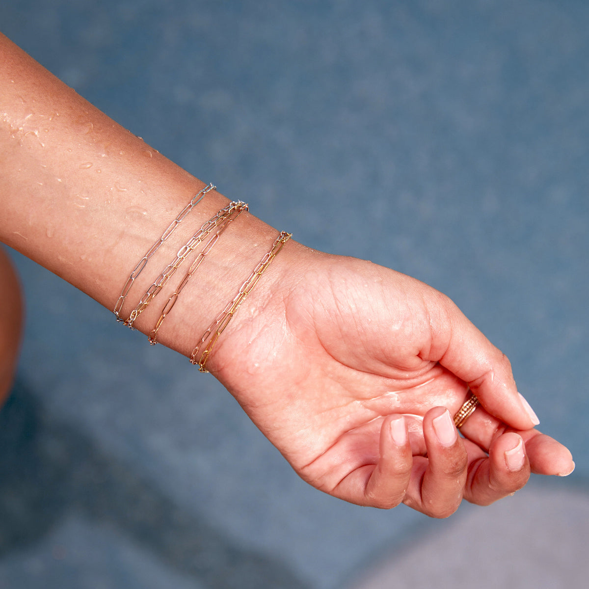 A hand wearing multiple mixed metal Element Long Link Bracelets and Element Short Link Bracelets and stacked gold rings, against a blue background.