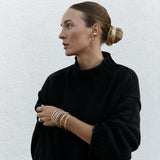 A woman wearing multiple stacked Flirt Bangles in gold and silver, a white gold Etched Paris Ring, an Open Diamond Ring and a C’est Moi Link Ring, two Ready to Mingle Ear Cuffs, Sleek & Chic Hoops and an Oval Hoop, all earrings in yellow gold, styled with her hair in a bun and a black knit.