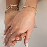 Two hands wearing several bracelets and rings in yellow or white gold: a Bold Link Bracelet, an Element Long Link Bracelet, two BFF Bracelets, a Down the Line Bracelet, a Link Ring and an Open Diamond Ring, against a neutral background. 