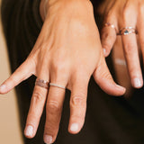 A close-up of a hand wearing rose gold Delicate Trio Stacking Rings and two Pavé Diamond Rings, with a blurred background showing another hand wearing multiple rings.