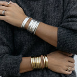 A close-up of arms wearing stacked Rendezvous Cuffs, Plus One Cuffs and All In Cuffs, one arm in silver and the other in gold, a Perla Pinky Ring, an Etched Paris Ring, an C'est Moi Link Ring and an Open Diamond Ring, styled in a dark grey knit.