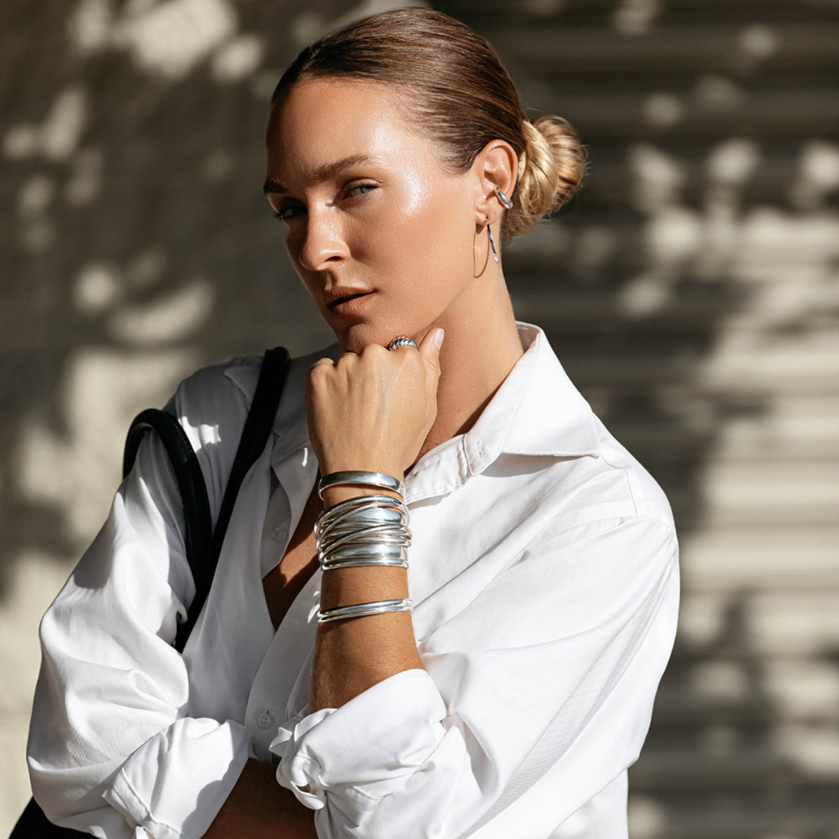 A woman wearing stacked sterling silver All In Cuffs and Plus One Cuffs, an Etched Paris Ring, a Ready to Mingle Ear Cuff and Sleek & Chic Hoops, with her hair in a bun and styled in a white shirt.