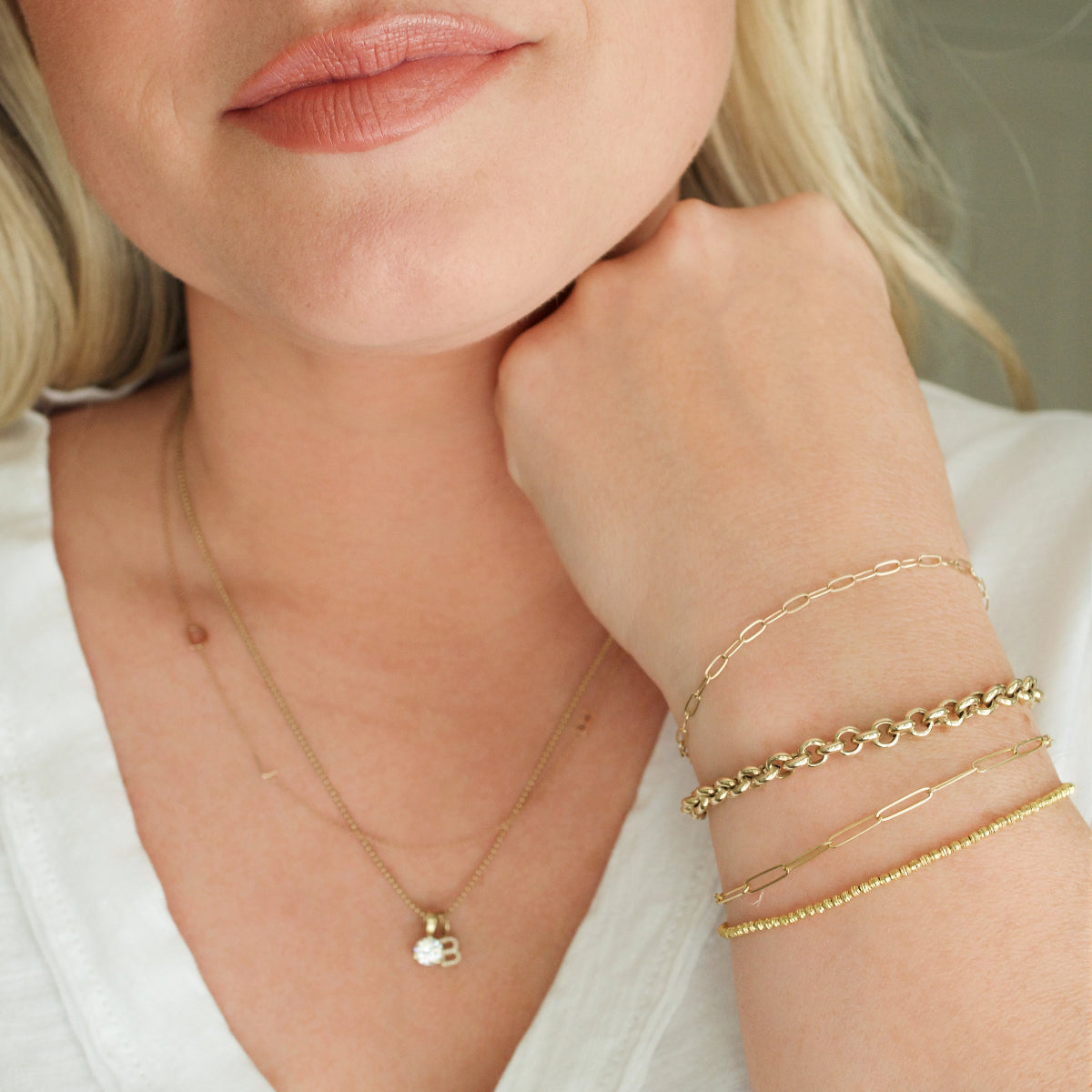 A close-up of a woman’s neck and wrist wearing an Element Short Link Bracelet, an Element Long Link Bracelet, a Rolo Bracelet and a Diamond Cut Bracelet, a Custom Gold Necklace with 2 letters (an L and a birthstone charm), and a Classic Bead Chain with a Diamond Letter Jumble featuring the letter B, all in yellow gold, styled with a white top.