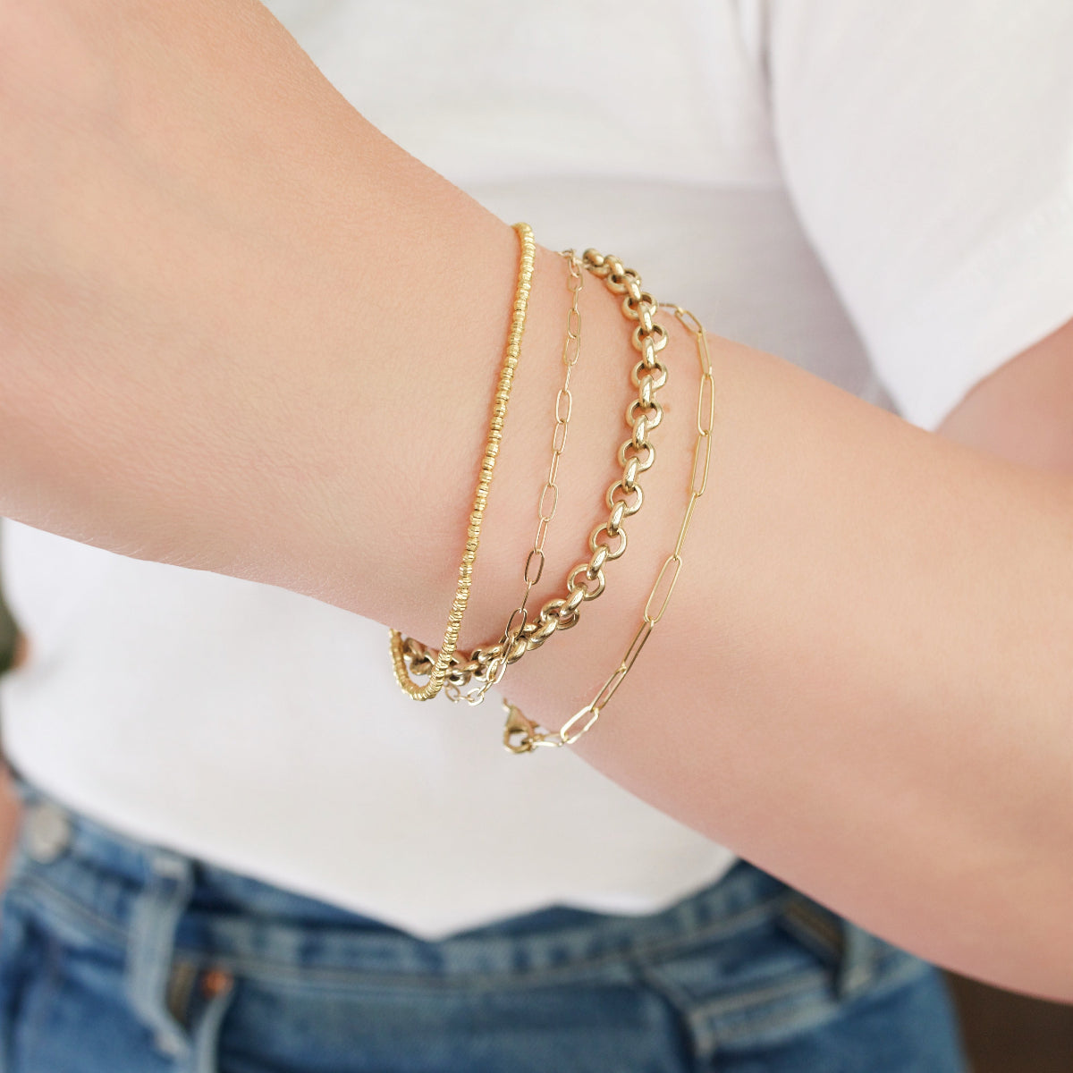 A close-up of a woman’s arm wearing yellow gold bracelets: a Diamond Cut Bracelet, an Element Long Link Bracelet, an Element Short Link Bracelet and a Rolo Bracelet, with a white top and blue jeans.