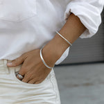 An arm wearing a silver Flirt Bangle and For Keeps Bangle and a white gold Etched Paris Ring, styled in a white shirt and off-white pants. 