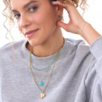 A woman wearing The Bluebell Bouquet Necklace, an Element Long Link Chain with a Turquoise Heart Charm, a Round Curb Chain with a Diamond Heart Locket Charm, a Circle Ear Jacket Pavé, a 14K Gold Pavé Letter Stud - C and a Birthstone Stud, with a light grey sweater and a white background. 