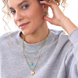 A woman wearing The Bluebell Bouquet Necklace, an Element Long Link Chain with a Turquoise Heart Charm, a Round Curb Chain with a Diamond Heart Locket Charm, a Circle Ear Jacket Pavé, a 14K Gold Pavé Letter Stud - C and a Birthstone Stud, with a light grey sweater and a white background. 