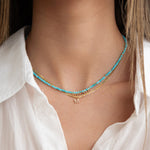 A close-up of a neck wearing an Endless Summer Necklace - Turqs + Caicos, an Overboard Layering Chain and a Pavé Letter Charm in M, styled in a white shirt. 