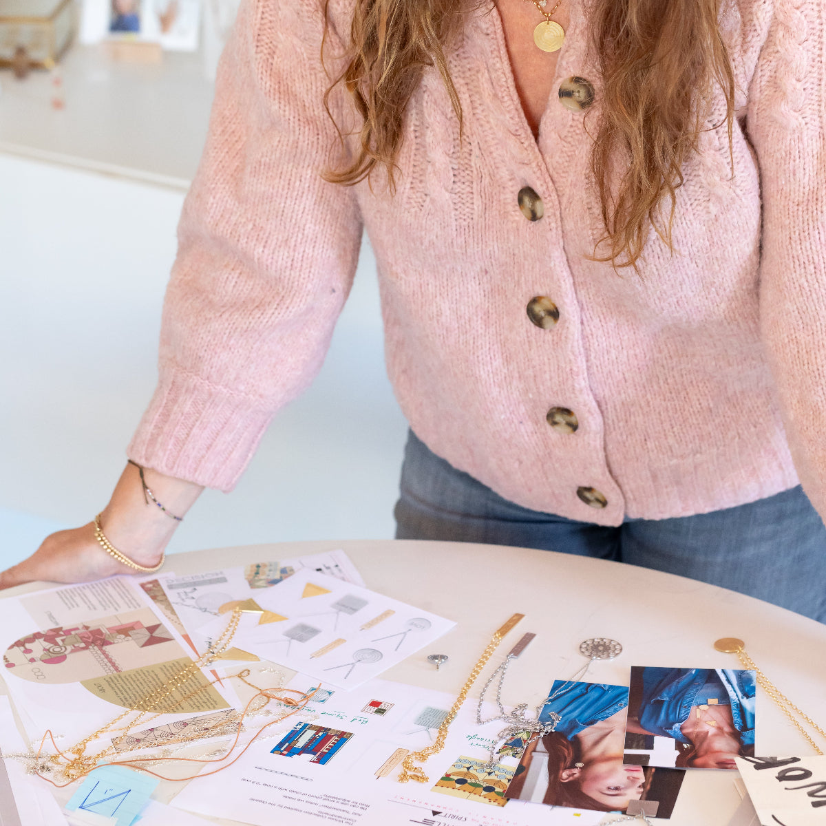 A woman wearing a Virtues Circle Pendant and Bubble Bracelets in yellow gold, styled with a light pink jumper and blue jeans, standing in front of a table displaying Virtues Pendants and a Skylight Necklace, and assorted papers and images.