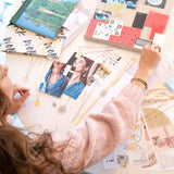 A woman working on jewelry design with Virtues Pendants - Circle and Rectangle in yellow gold and sterling silver, a Skylight Necklace and Hoop in sterling silver. laid out with photos and books. She is wearing Bubble Bracelets. Geo Stacking Rings and a light pink jumper. 