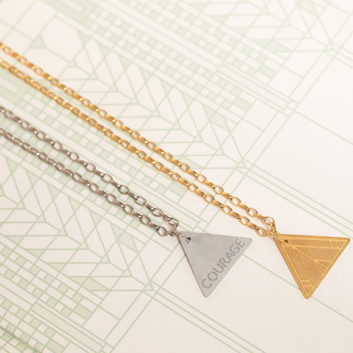 Two Virtues Pendants - Triangle, one in yellow gold and the other in sterling silver  on a geometric patterned background. 