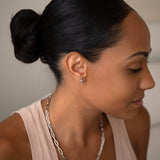 A woman wearing a white gold Preppy Post and a Bold Link Chain with a nude tank top and her black hair in a bun, against a light grey background.