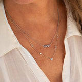 A close-up of a woman's décolletage wearing a NANA Necklace and a Custom Gold Necklace - 5 Letters, both in white gold with beaded chains, paired with a white shirt.