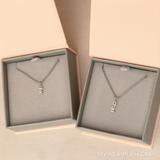 Two white gold Box Chain necklaces each with a Petite Nameplate Pendant - one reading ‘dixie’ and the other ‘charli’, placed in separate light grey and light pink jewellery boxes on a beige background.