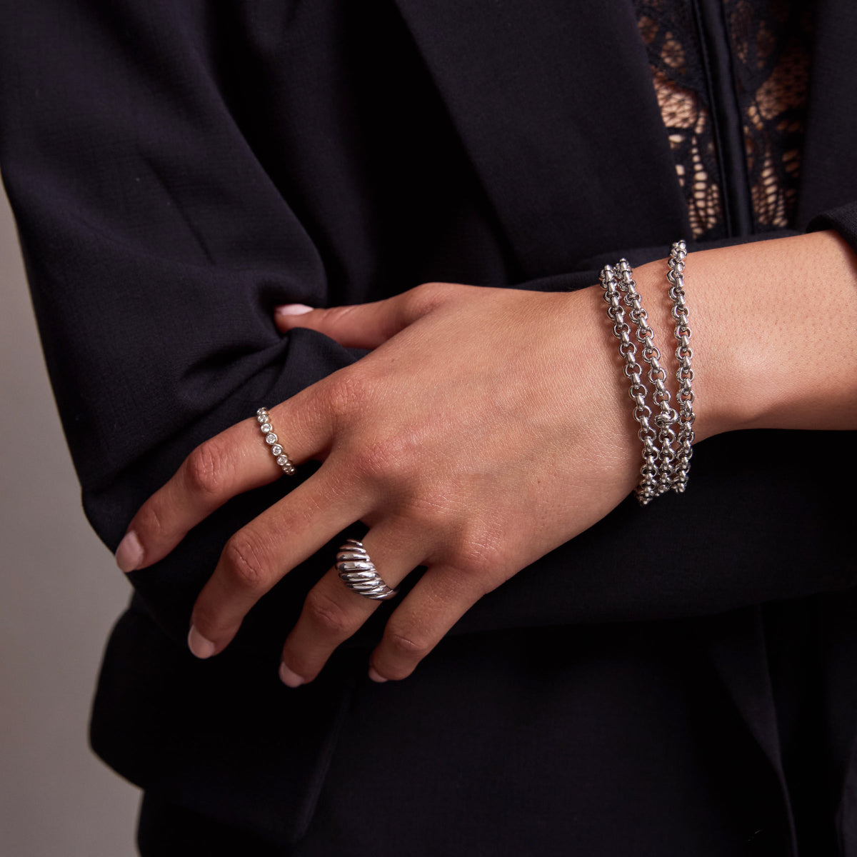 A close-up of a woman’s hand wearing three Rolo Bracelets, an Evermore Ring and an Etched Paris Ring, all in white gold, with a navy suit and lace top.