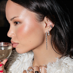 The Night Cap Ear Cuff, a Triple Sec Diamond Earring, Diamond Cascade Earring and Love on the Rocks Ring being worn by a brunette woman with a cream fluffy jacket holding a cocktail.