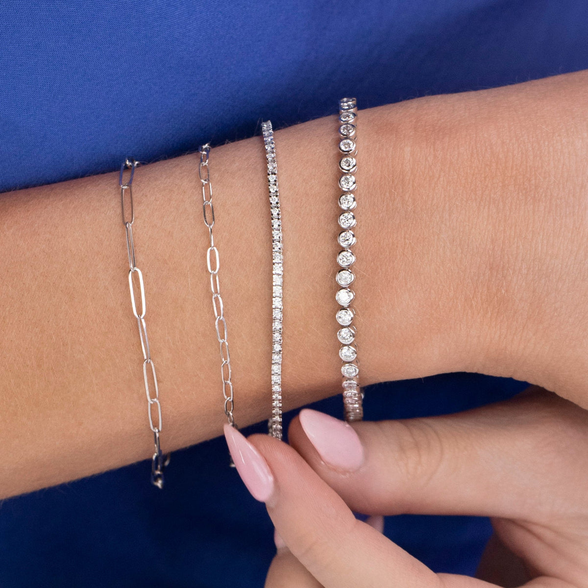 A close-up of a wrist wearing an Element Short Link Bracelet, an Element Long Link Bracelet, a Down the Line Bracelet and a Love Set Match Bracelet, against a blue background.