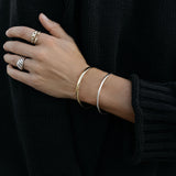 A close-up of an arm wearing a gold For Keeps Bangle and silver Flirt Bangle, a white gold Etched Paris Ring, an Open Diamond Ring and a C’est Moi Link Ring, styled with a black knit. 