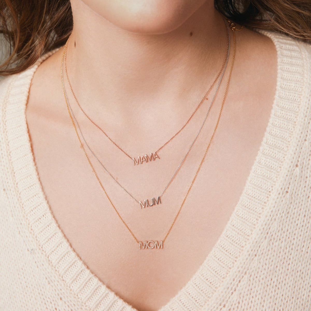 A close-up of a woman's décolletage wearing a MAMA Necklace in rose gold, a MUM Necklace in white gold and a Pavé MOM Necklace in yellow gold, with a cream knit top and brown hair. 