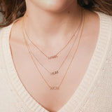 A close-up of a woman's décolletage wearing a MAMA Necklace in rose gold, a MUM Necklace in white gold and a Pavé MOM Necklace in yellow gold, with a cream knit top and brown hair. 