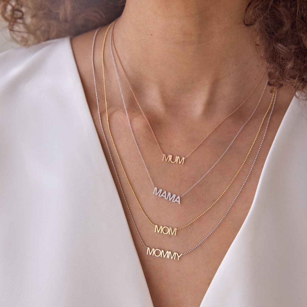 Close-up of a woman's décolletage wearing a MUM Necklace in rose gold, a Pavé MAMA Necklace in white gold, a MOM Necklace in yellow gold and a MOMMY Necklace in mixed metal, with a white top and brown curly hair. 