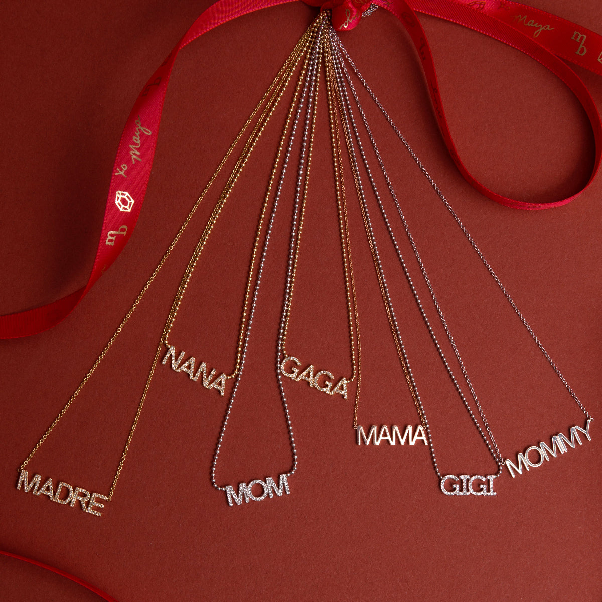 Pavé MADRE, Pavé NANA, Pavé GAGA, Pavé GIGI, MAMA, and MOMMY Necklaces tied to a red ribbon against a red background.
