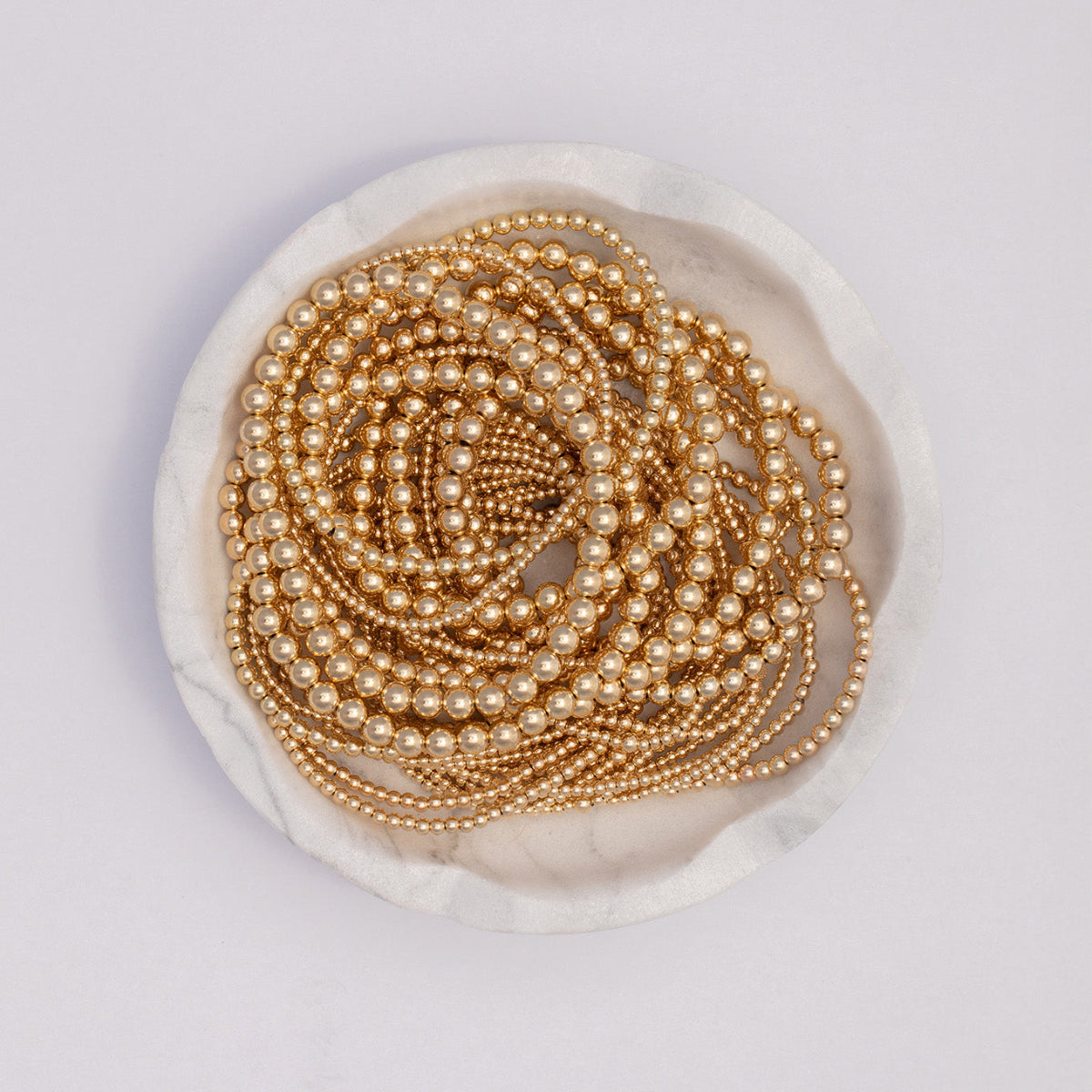 Yellow gold Bubble Bracelets in various sizes coiled in a white marble bowl, against a white background.