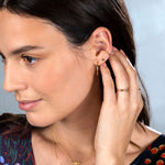 A Birthstone Stud, Rounded Ear Cuff, yellow gold hoop earring, two Birthstone Rings and LOVE Necklace worn by a brunette woman with a floral navy top against a light blue background. 